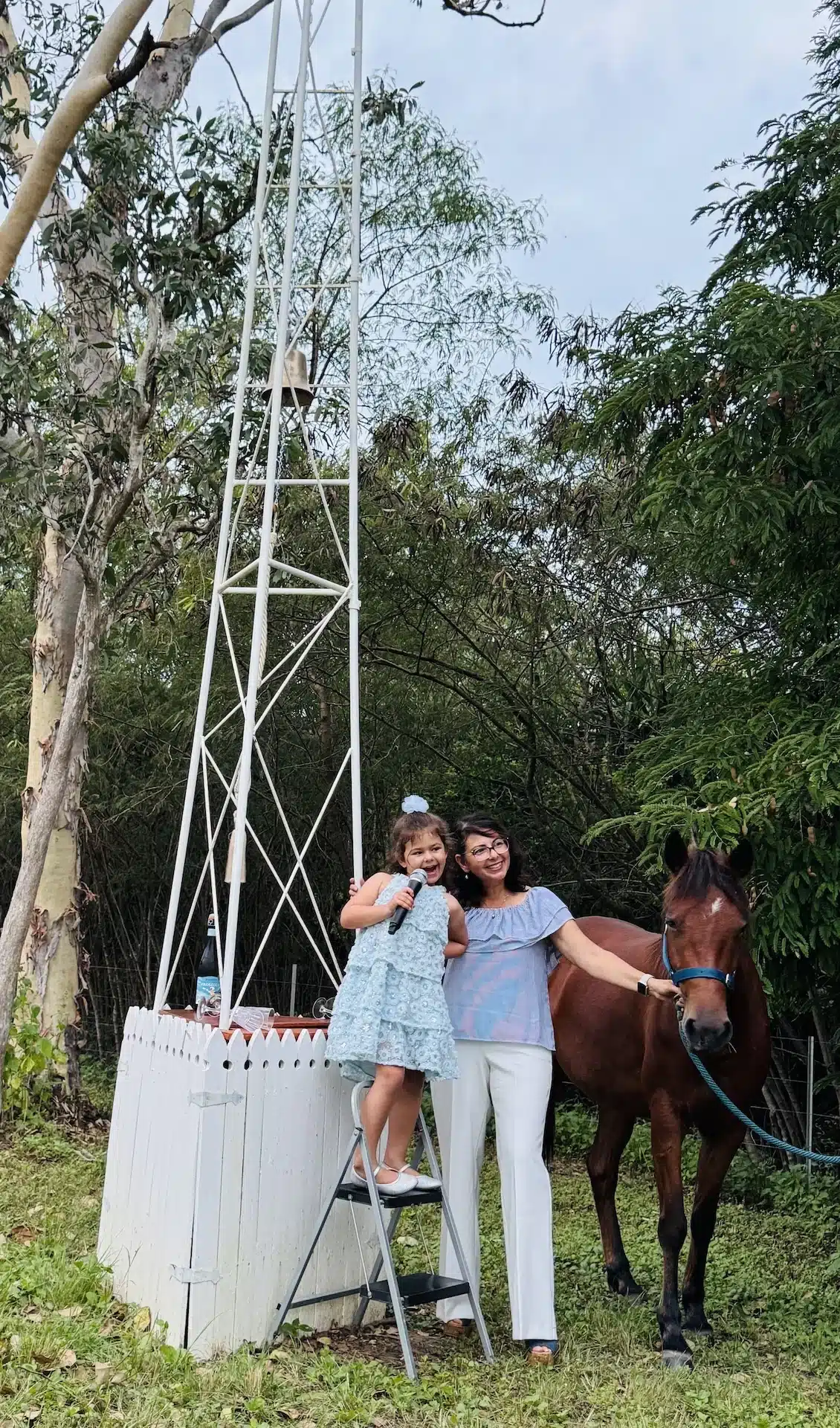 IMG_7994 A young girl in a light blue dress sings into a microphone while standing on a step ladder next to a white metal tower, with a smiling woman and a brown horse beside them in a grassy yard.