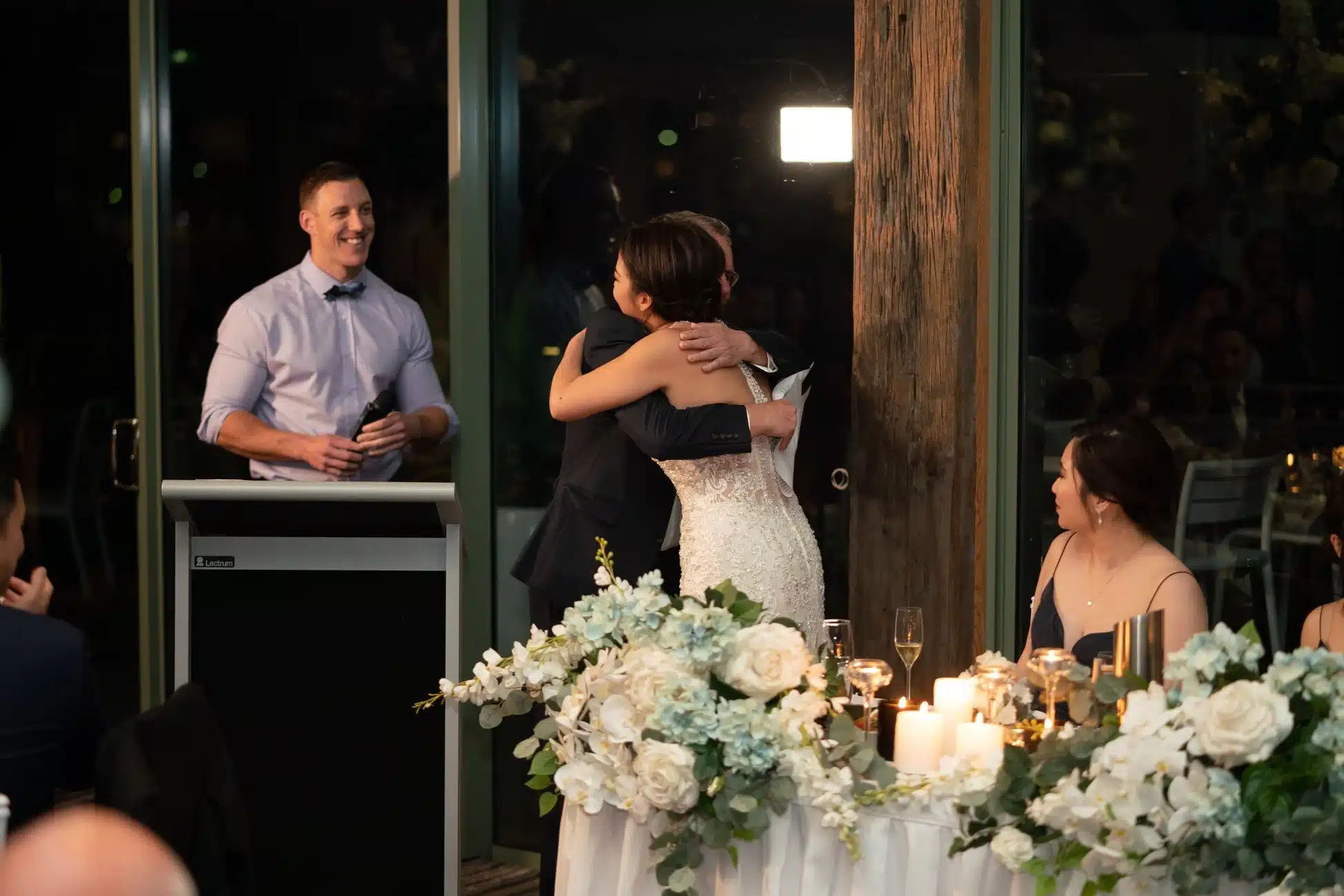 Image 6 Bride in a lace wedding gown hugs a man in a dark suit at a reception, candles and floral centerpiece in foreground.