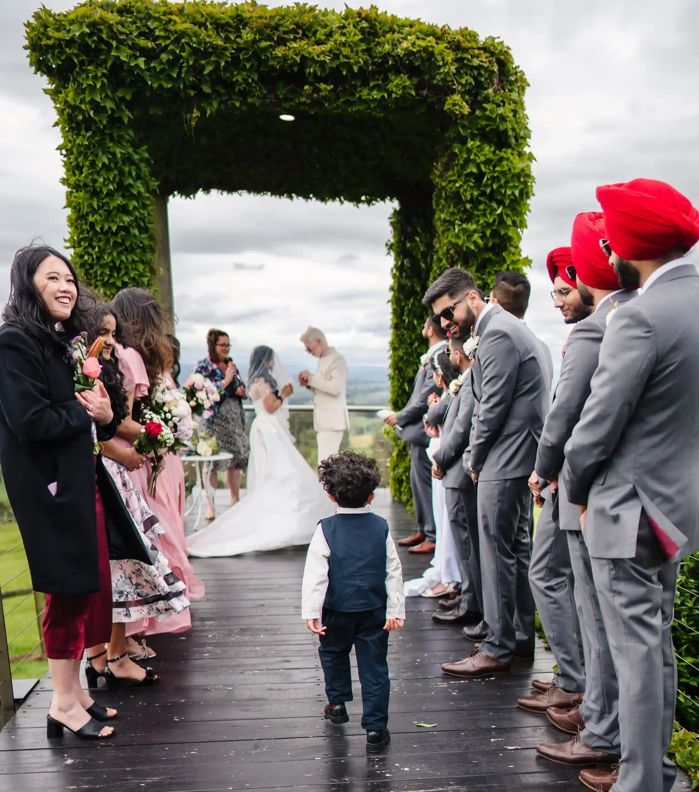 Andrea & Robin Young boy in a navy vest walks down the wooden aisle toward the wedding couple, flanked by bridesmaids and groomsmen in gray.