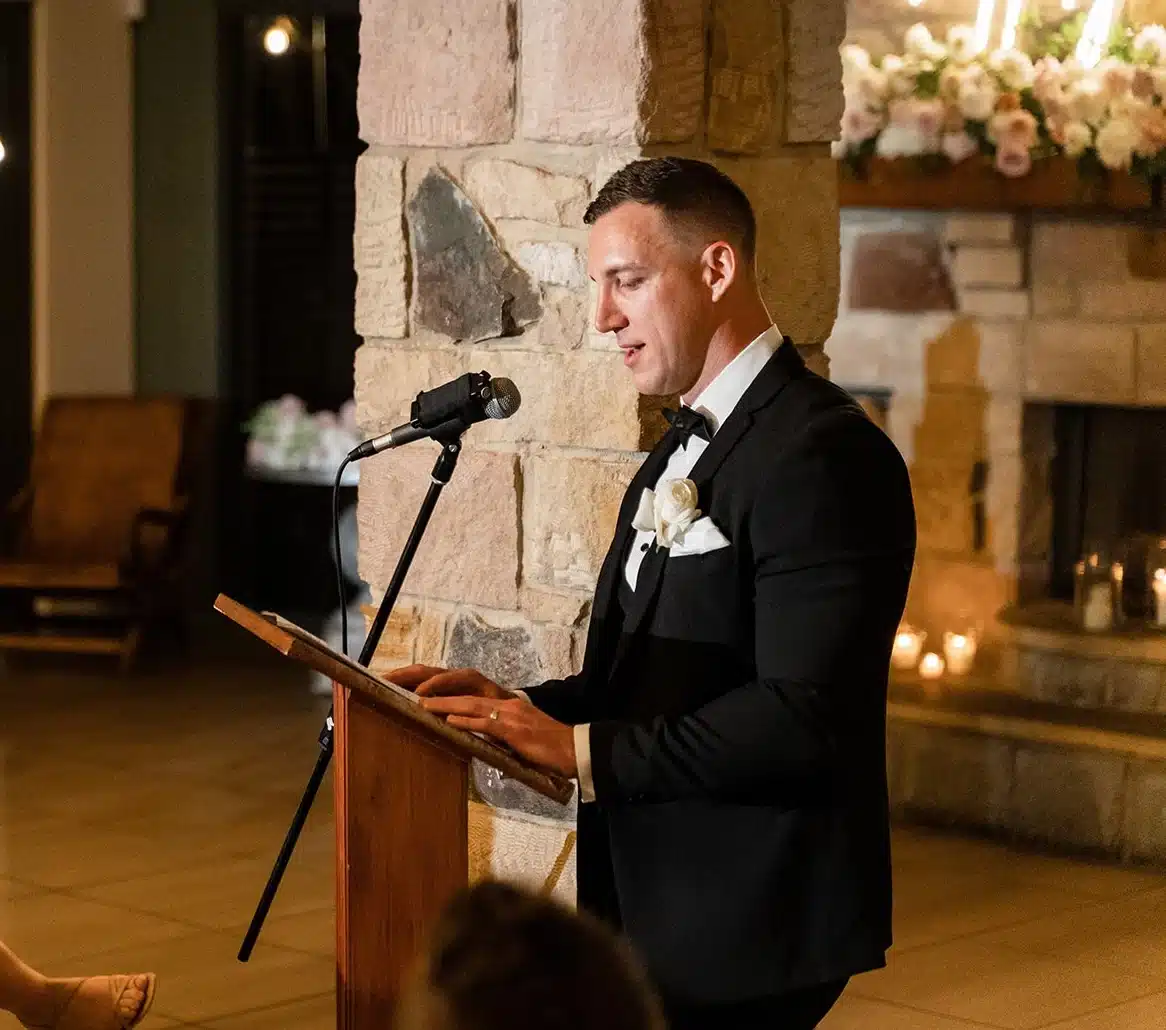 Sydney Mc Man in a black tuxedo delivering a speech at a wooden podium with a microphone in a stone-walled venue.