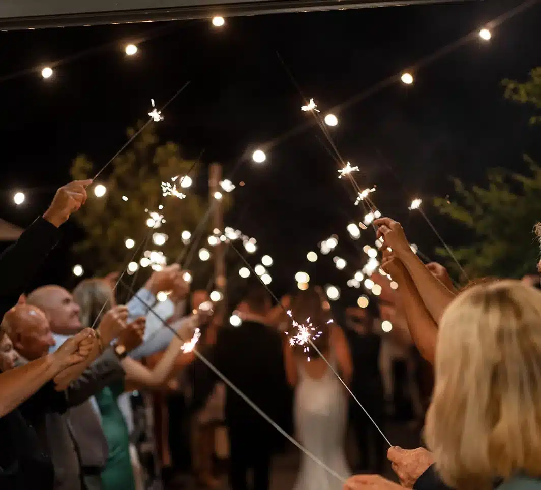 WD221202P2 AlexandraRiley J1494-912 copy Group of people at night holding lit sparklers, creating a festive line of sparks under string lights.