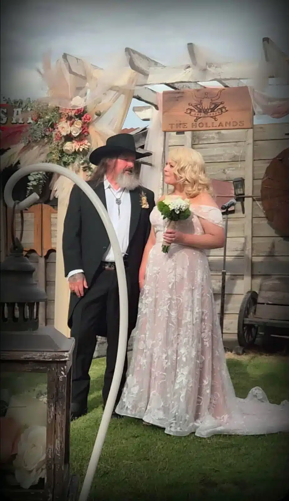 attachment-weddingjpeg A bearded man in a black suit and cowboy hat stands beside a bride in a lace gown holding a bouquet at a rustic outdoor wedding backdrop.
