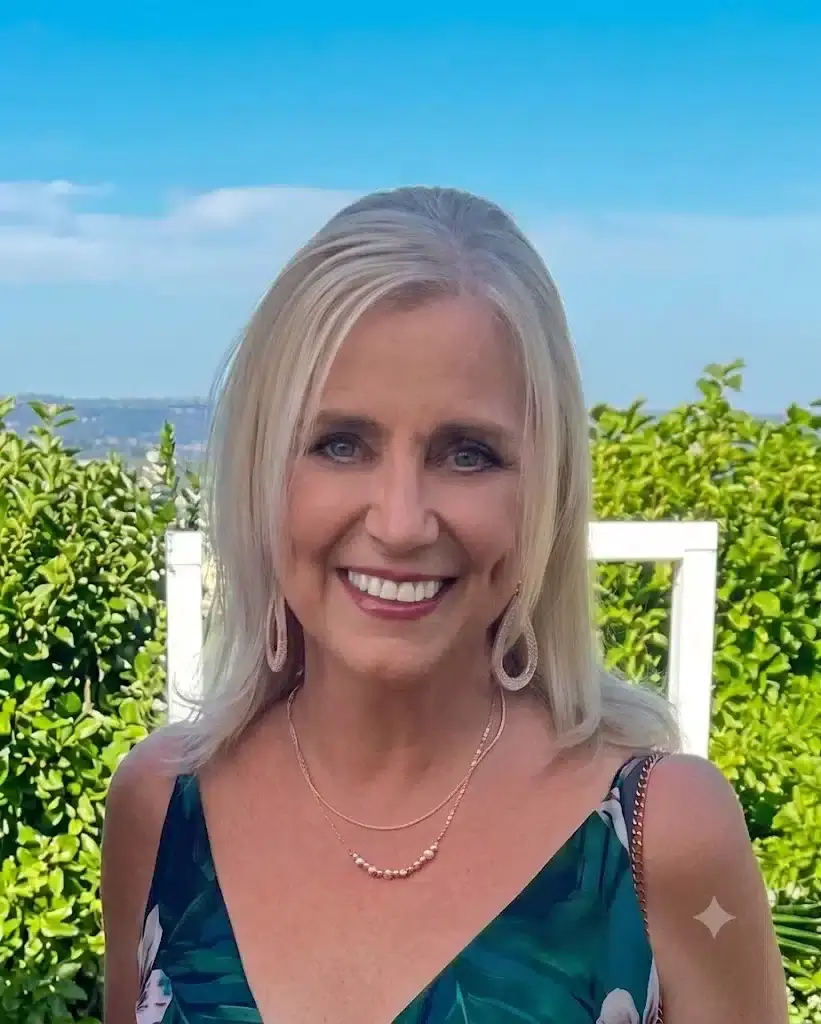 Fiona Da Silva Celebrant 1 Smiling woman with blonde hair in a teal green dress outdoors, blue sky and shrubbery behind her.