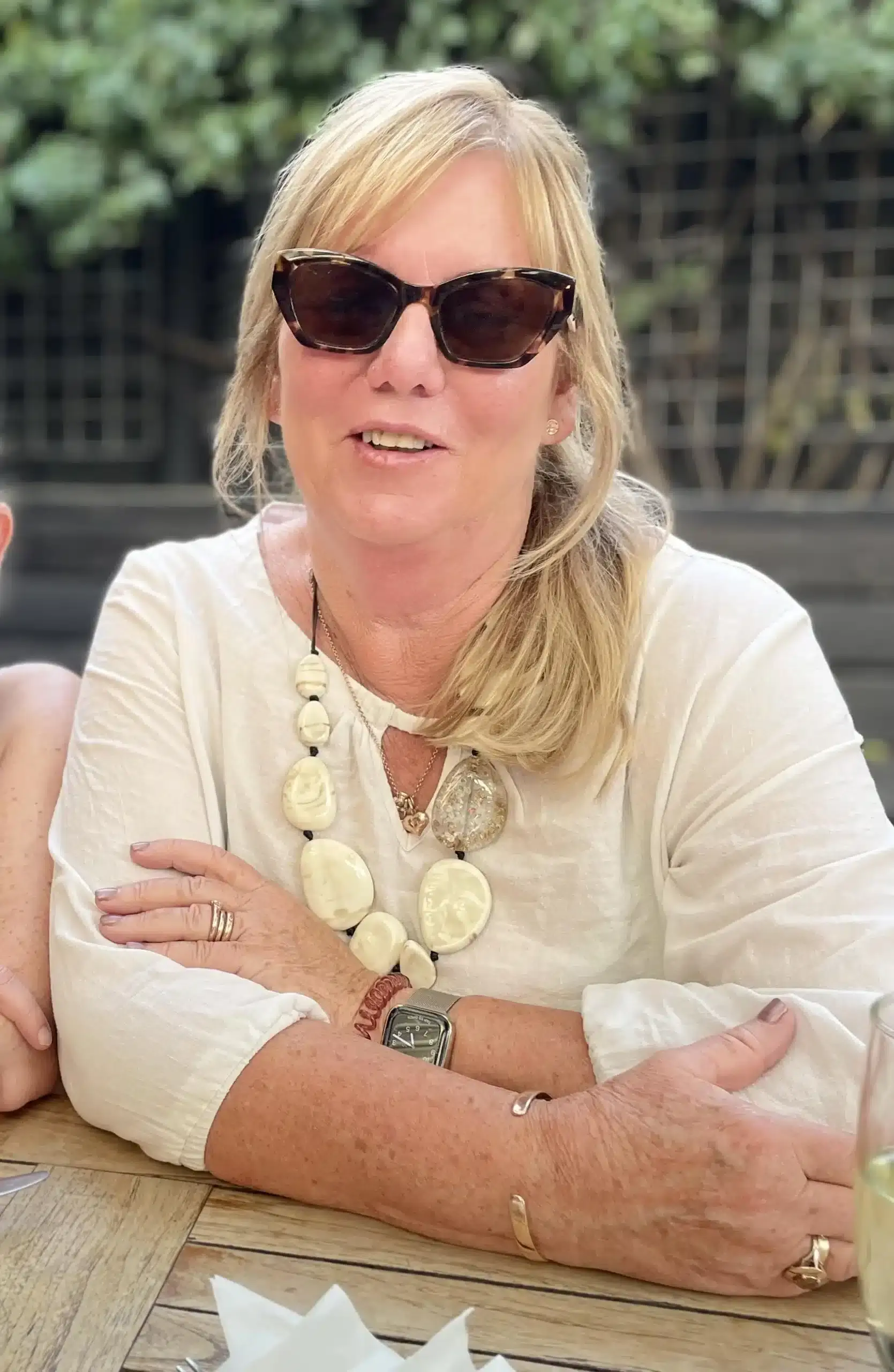 louise-celebrantjpg Older woman with blonde hair wearing dark tortoiseshell sunglasses and a cream necklace sits at a wooden table outdoors, arms folded.