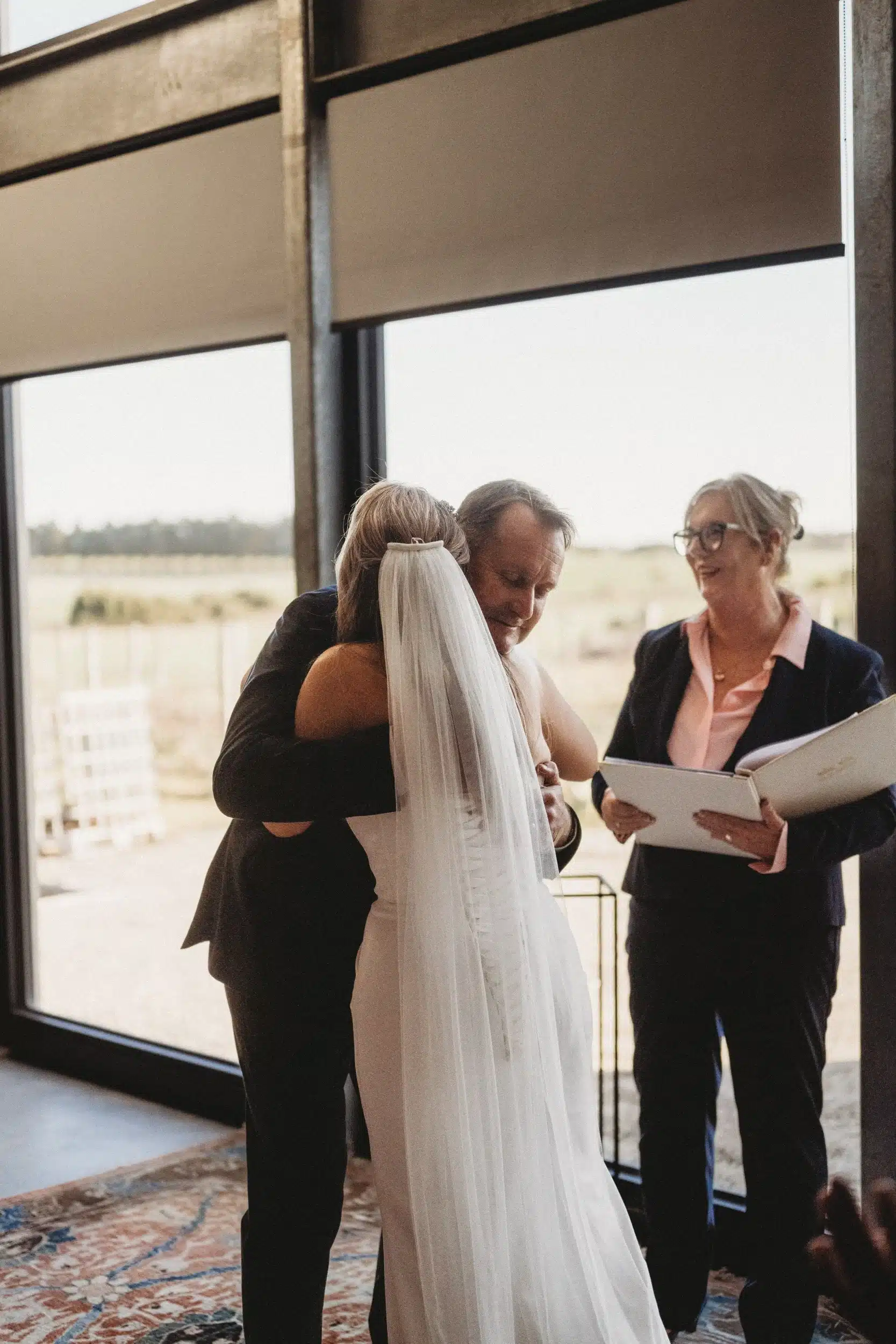 wedding-photos-m-d-3-minjpg Bride in a white gown with a veil embraces a partner during a wedding ceremony as the officiant reads from a folder nearby.